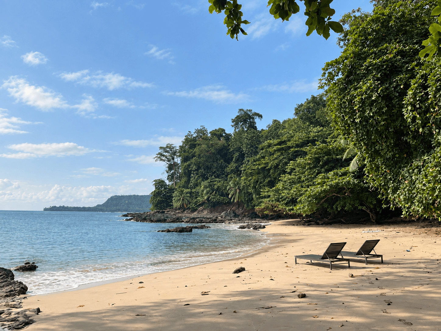 Uma praia dourada com rochas negras e floresta tropical até à água, e duas espreguiçadeiras na areia.
